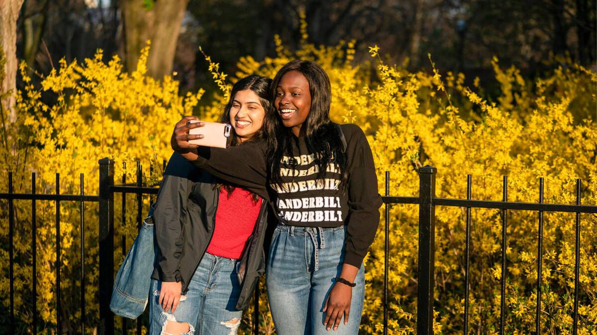 two female students take a selfie in front of bright yellow flowers on campus