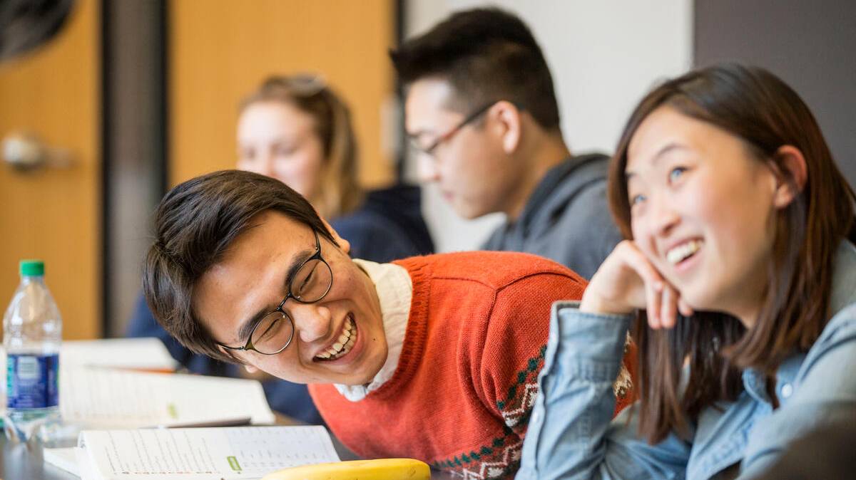 a student sitting at a table leans forward and laughs, looking to his left and other students seated at the table