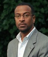 Headshot of Paul Taylor, a young black man standing outside in front of a vine covered wall. He is wearing a gray suit and a white shirt.